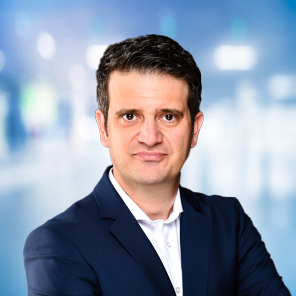 Mann in Anzug mit verschränkten Armen, vor einem unscharfen, modernen Hintergrund.