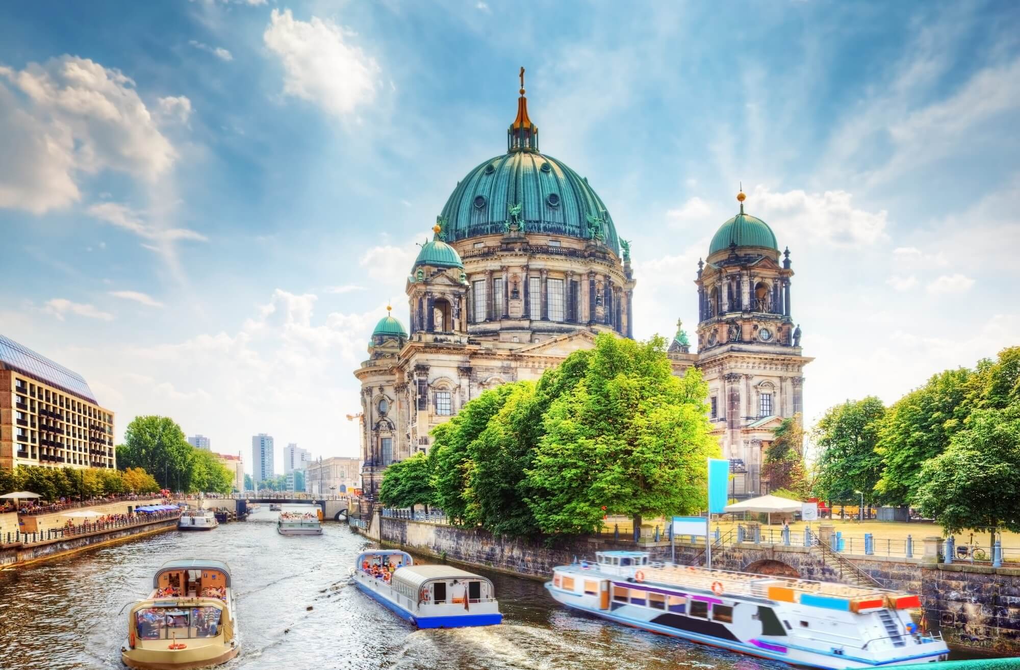 Berliner Dom mit gr&uuml;nen Kuppeln, umgeben von B&auml;umen und Wasserwegen, an einem sonnigen Tag.