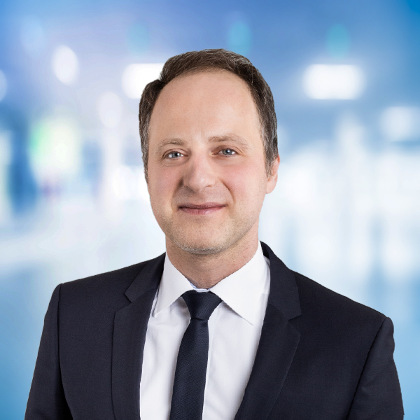Ein lächelnder Mann in einem Anzug vor einem unscharfen, hellen Hintergrund.