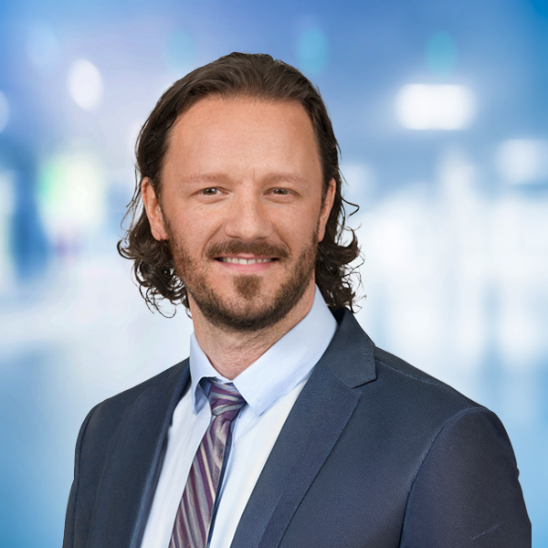 Mann mit lockigem Haar, Anzug und Krawatte, lächelt vor unscharfem, hell erleuchtetem Hintergrund.