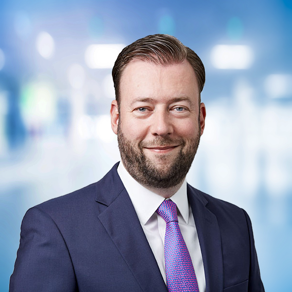 Mann in Anzug mit blauer Krawatte, lächelnd vor unscharfem, modernem Hintergrund.