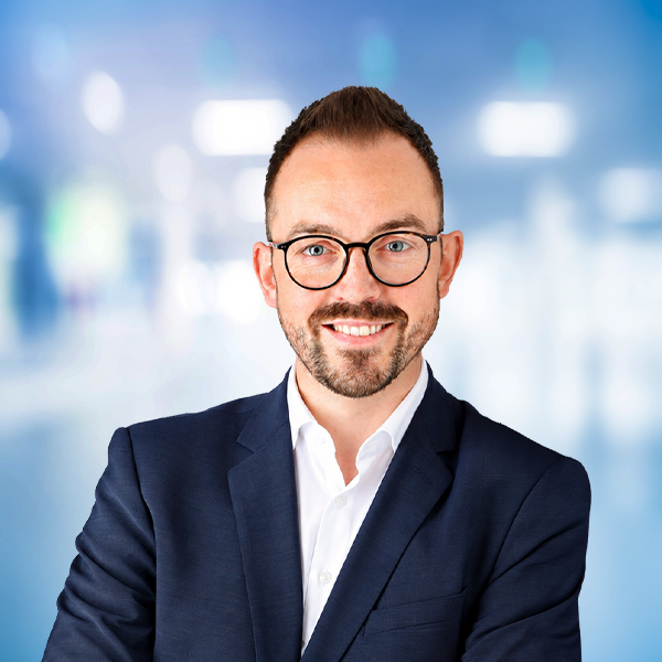 Ein lächelnder Mann mit Brille trägt einen Anzug und steht vor einem unscharfen, hellen Hintergrund.