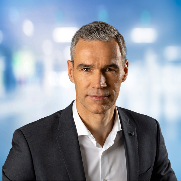 Porträt eines Mannes in einem Anzug mit grauen Haaren, vor einem unscharfen, modernen Hintergrund.