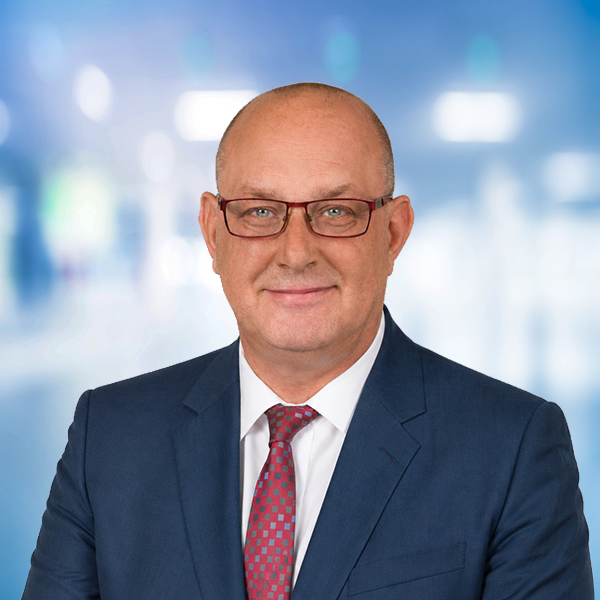 Mann mit Brille und Anzug, lächelnd vor unscharfem Hintergrund in einem modernen Umfeld.
