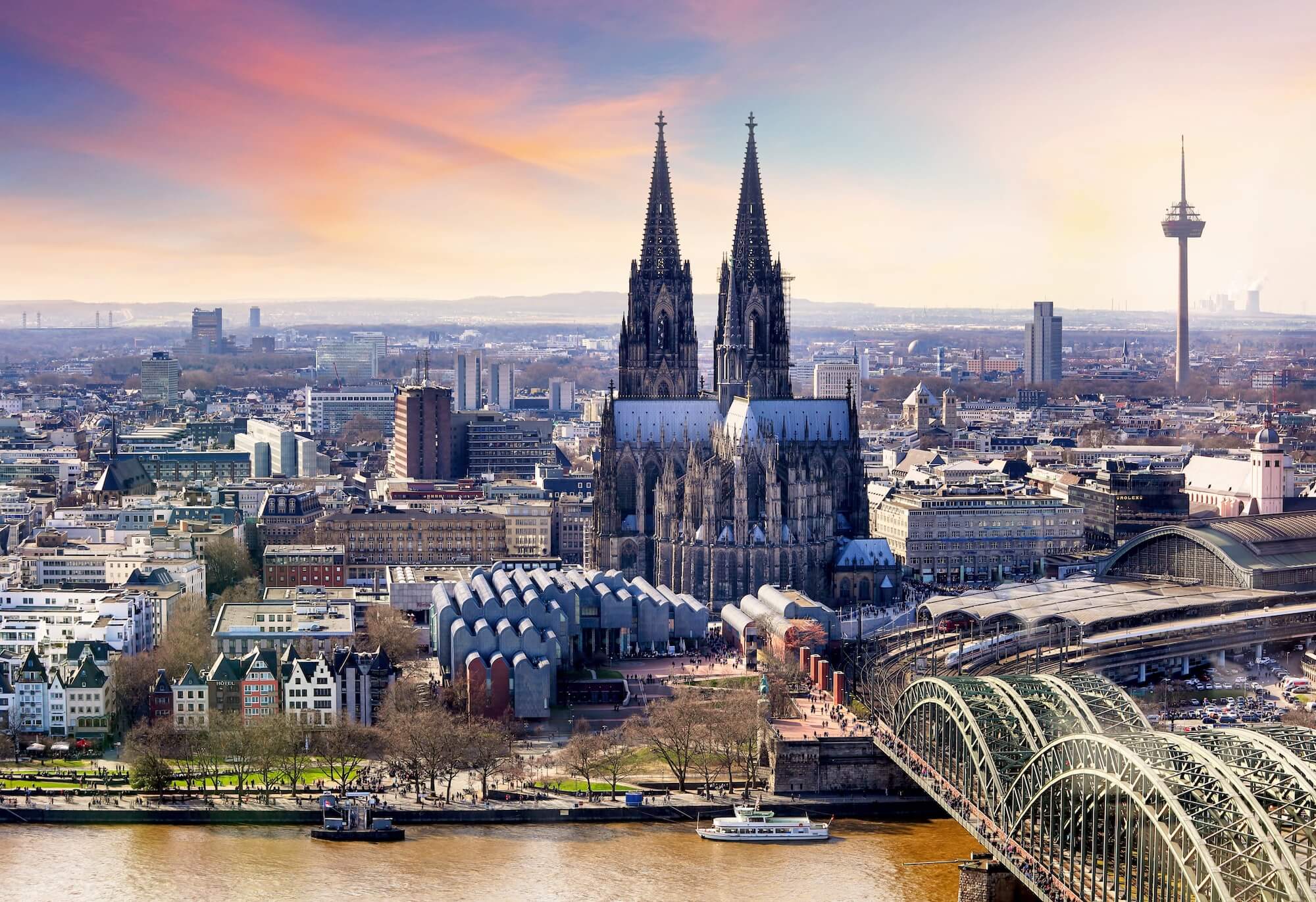 K&ouml;lner Dom mit zwei T&uuml;rmen im Vordergrund, umgeben von Stadtlandschaft und Rhein, bei Sonnenuntergang.