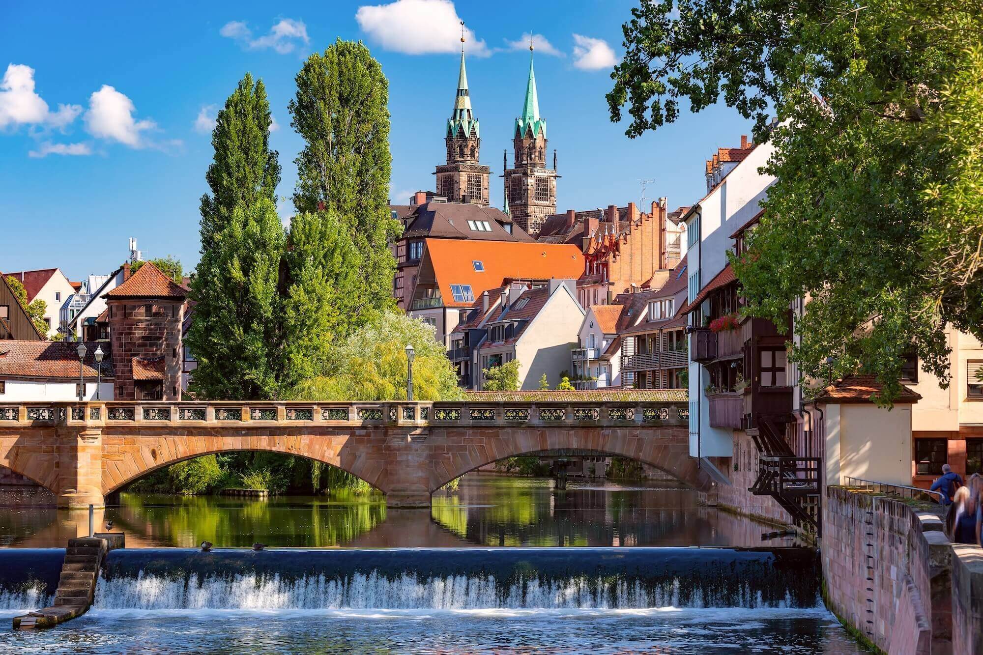 Blick auf eine Brücke über einen Fluss mit historischen Gebäuden und zwei Türmen im Hintergrund.