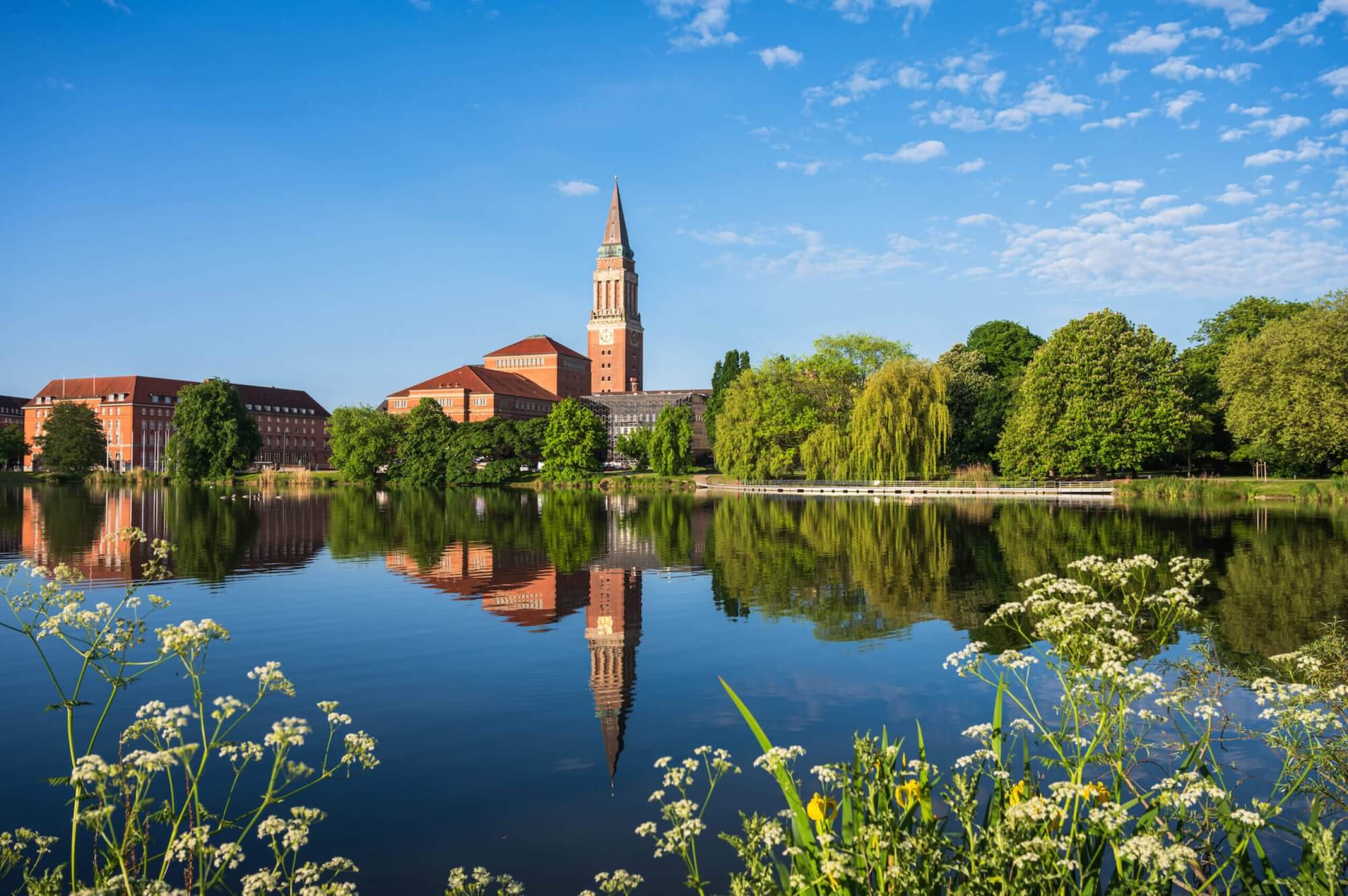 Ansicht des Rathauses von Kiel am Ufer eines ruhigen Sees, umgeben von Bäumen und blühenden Pflanzen.