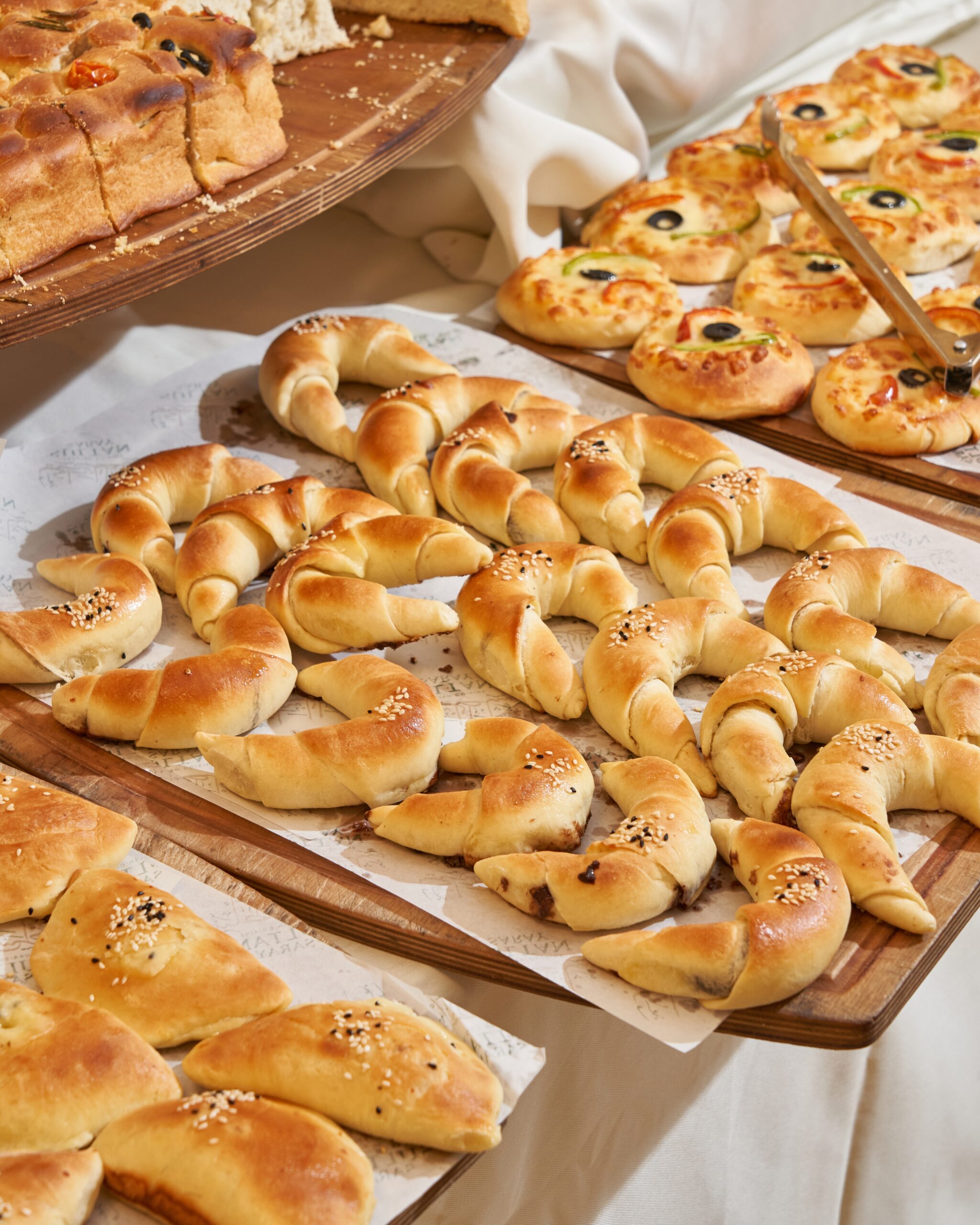 Ein Buffet mit verschiedenen Brötchen und Gebäck, darunter gedrehte Brötchen und belegte Fladen, auf Holzplatten.