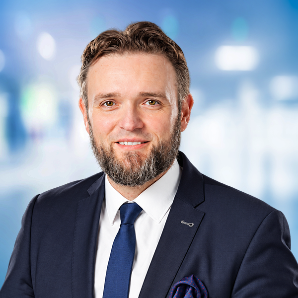 Ein Mann mit Bart in Anzug und Krawatte l&auml;chelt vor einem unscharfen, hellen Hintergrund.