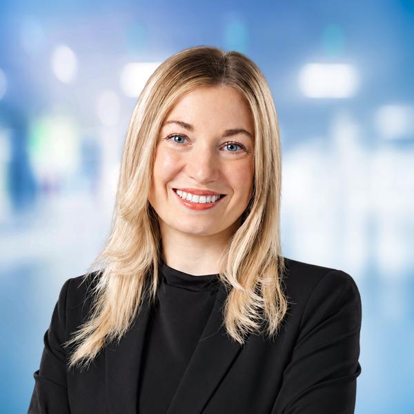 Eine l&auml;chelnde Frau mit blonden Haaren in einem schwarzen Blazer, vor einem unscharfen, hellen Hintergrund.
