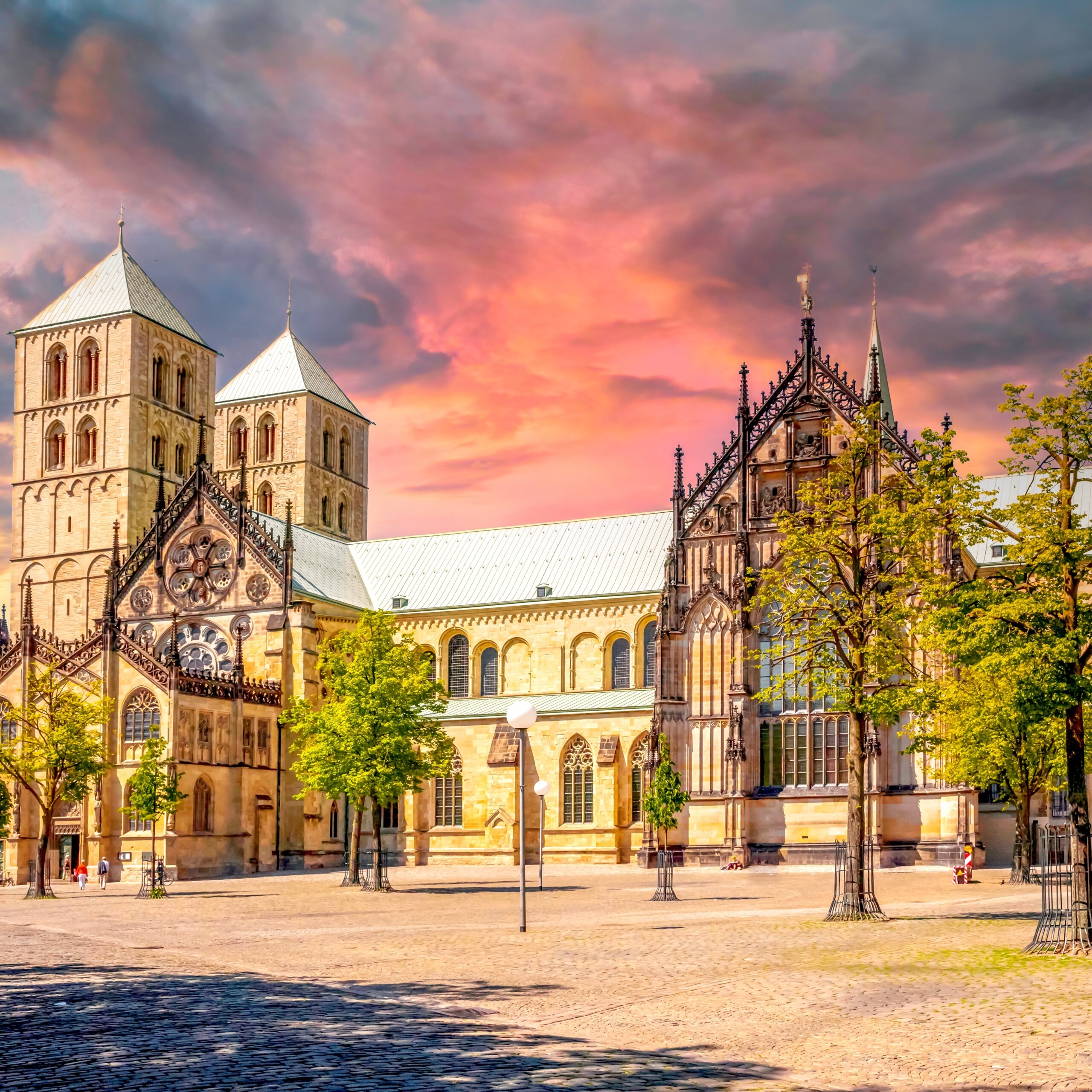 Historische Kathedrale mit T&uuml;rmen und buntem Himmel, umgeben von B&auml;umen und einem belebten Platz.